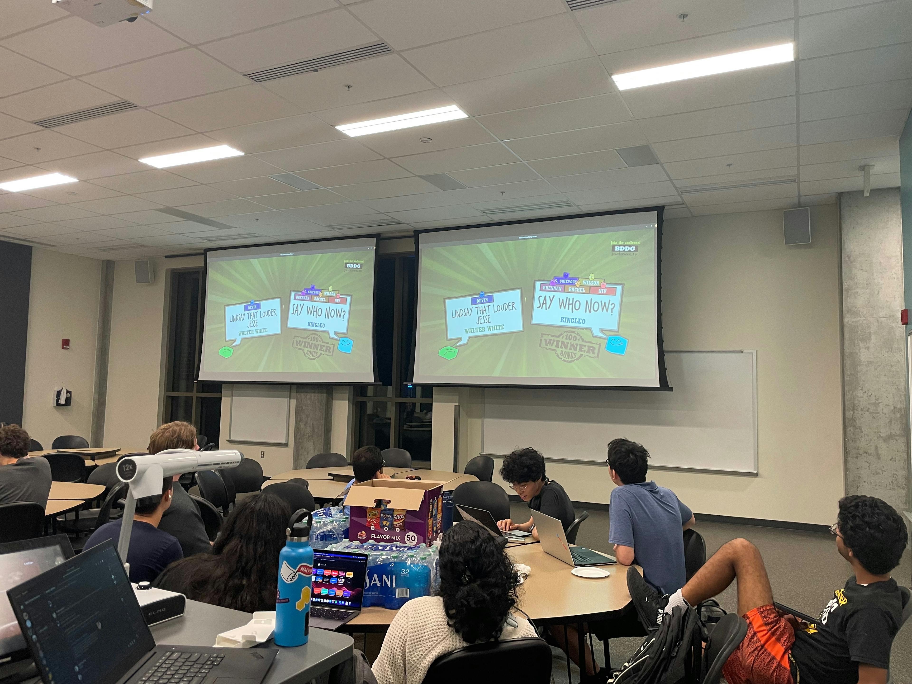 People sitting at tables in a large room, looking up at a projector screen which has a Jackbox Game playing